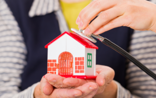 A senior woman holds a house in her hand as someone holds a stethoscope to it, symbolizing home care.