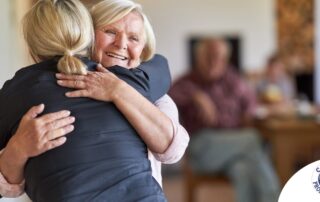 An older woman smiles as a younger woman visits her and hugs her, showing the effect that acts of kindness can have on senior loved ones.